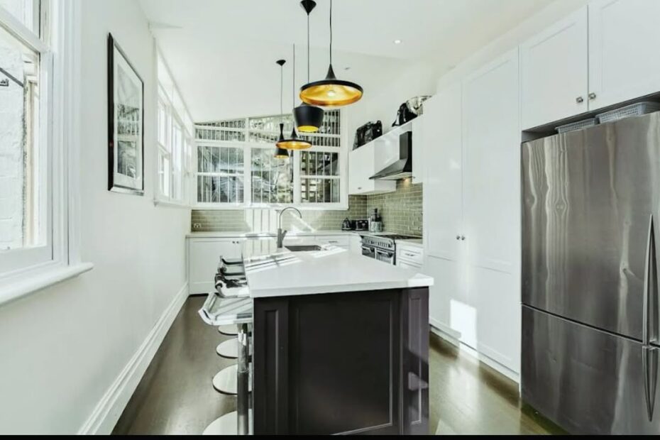 Modern kitchen interior with white shaker cabinets, a dark central island with a white countertop and bar stools, and three black and gold pendant lights hanging above.